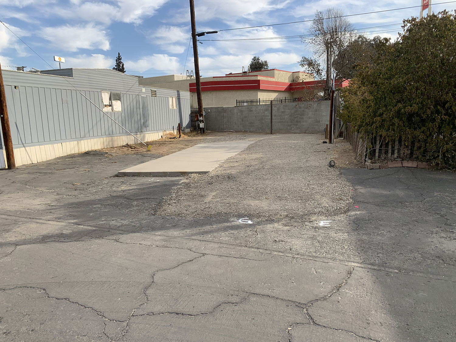 Empty RV parking space with a concrete pad and gravel area at Park View Mobile Home & RV Park in Fresno, CA, next to a gray mobile home and surrounded by a fence, trees and utility poles under a partly cloudy sky.
